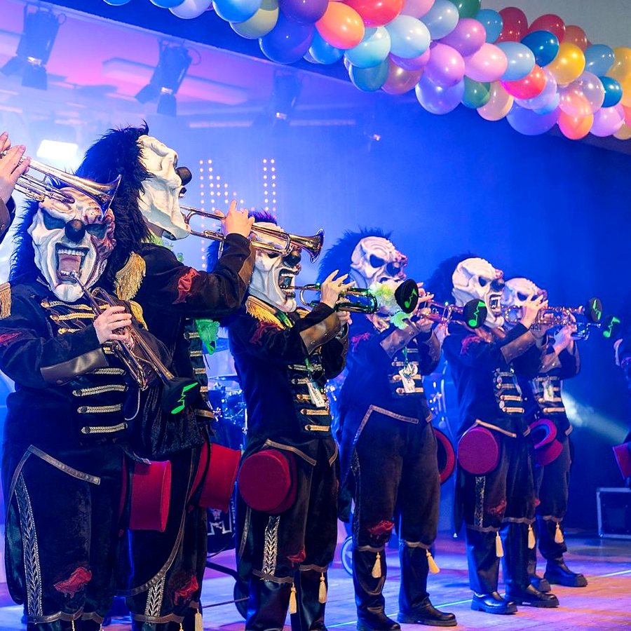 Die Lärmer auf der Bühne: eine Reihe Trompeten, Horror-Clown-Masken und schwarze Uniformjacken unter bunten Luftballons