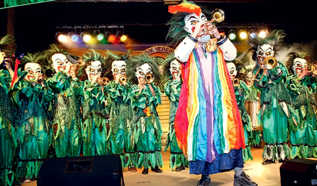Gugge in grünen Fransenkostümen mit weißen Masken, vorne ein Musiker im Regenbogenmantel mit Trompete
