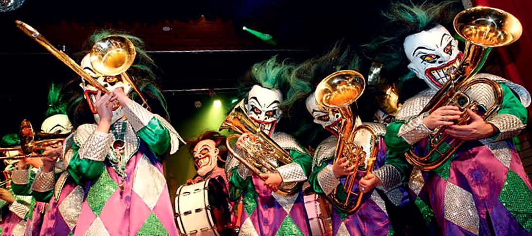 Gugge in Joker-Masken mit grünen Haaren und Rautenkostümen, mit Posaune, Sousaphon und Trommel
